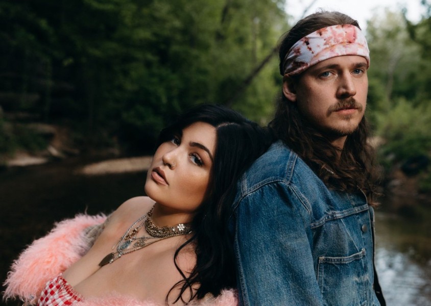 A man and woman stood back to back in front of a river. the woman is wearing a pink fluffy coat around her shoulders and lots of silver necklaces. The man has on a blue denim jacket and pink and white tie dye bandana around his headx